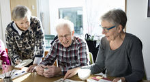 En ældre mand og to ældre kvinder sidder ved et bord og taler sammen over en avis